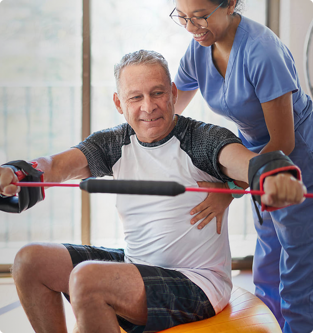Senior man exercising with resistance bands on an exercise ball, assisted by a smiling female therapist in scrubs.