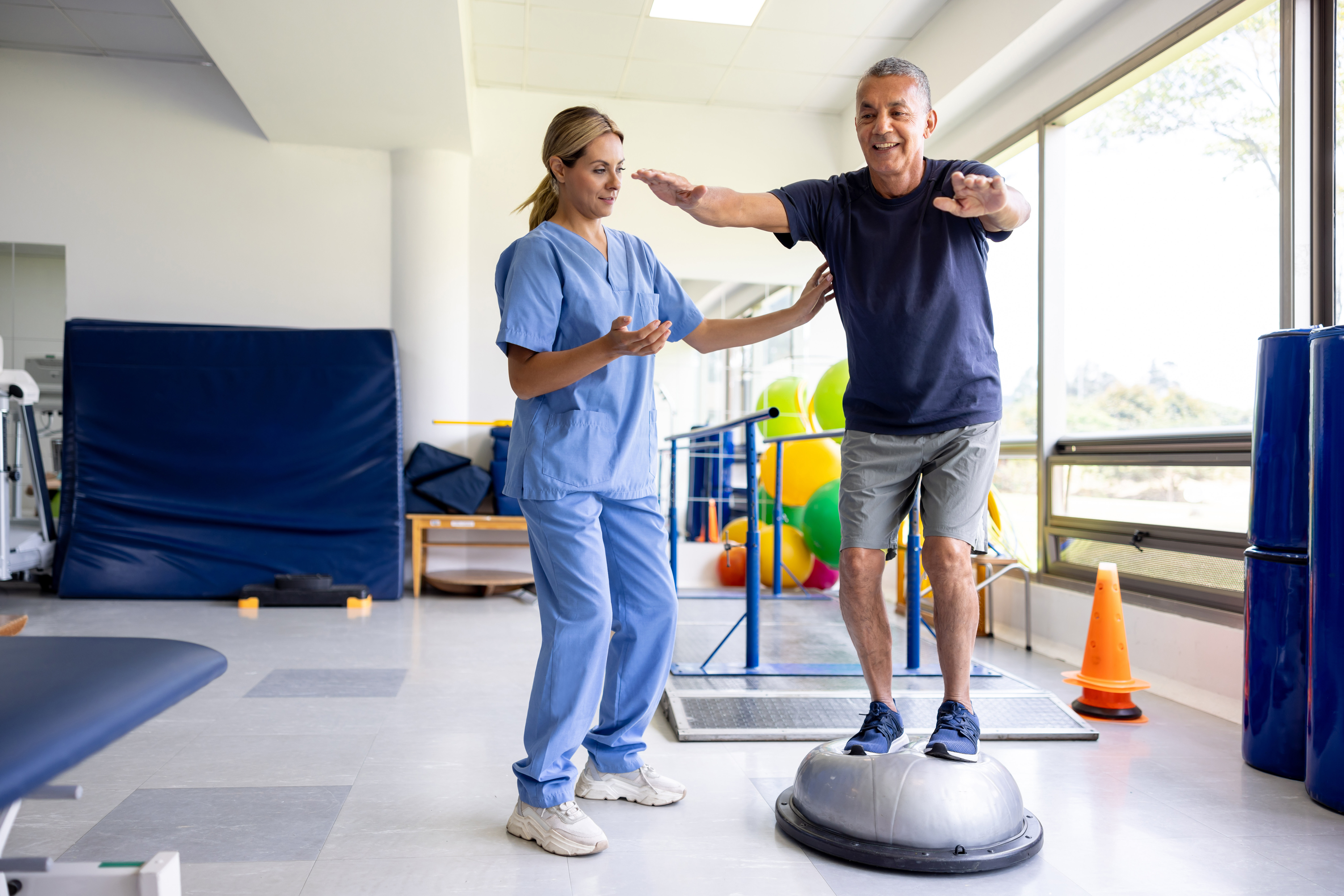 Physical therapist assisting a man balancing on a stability dome in a rehabilitation gym.