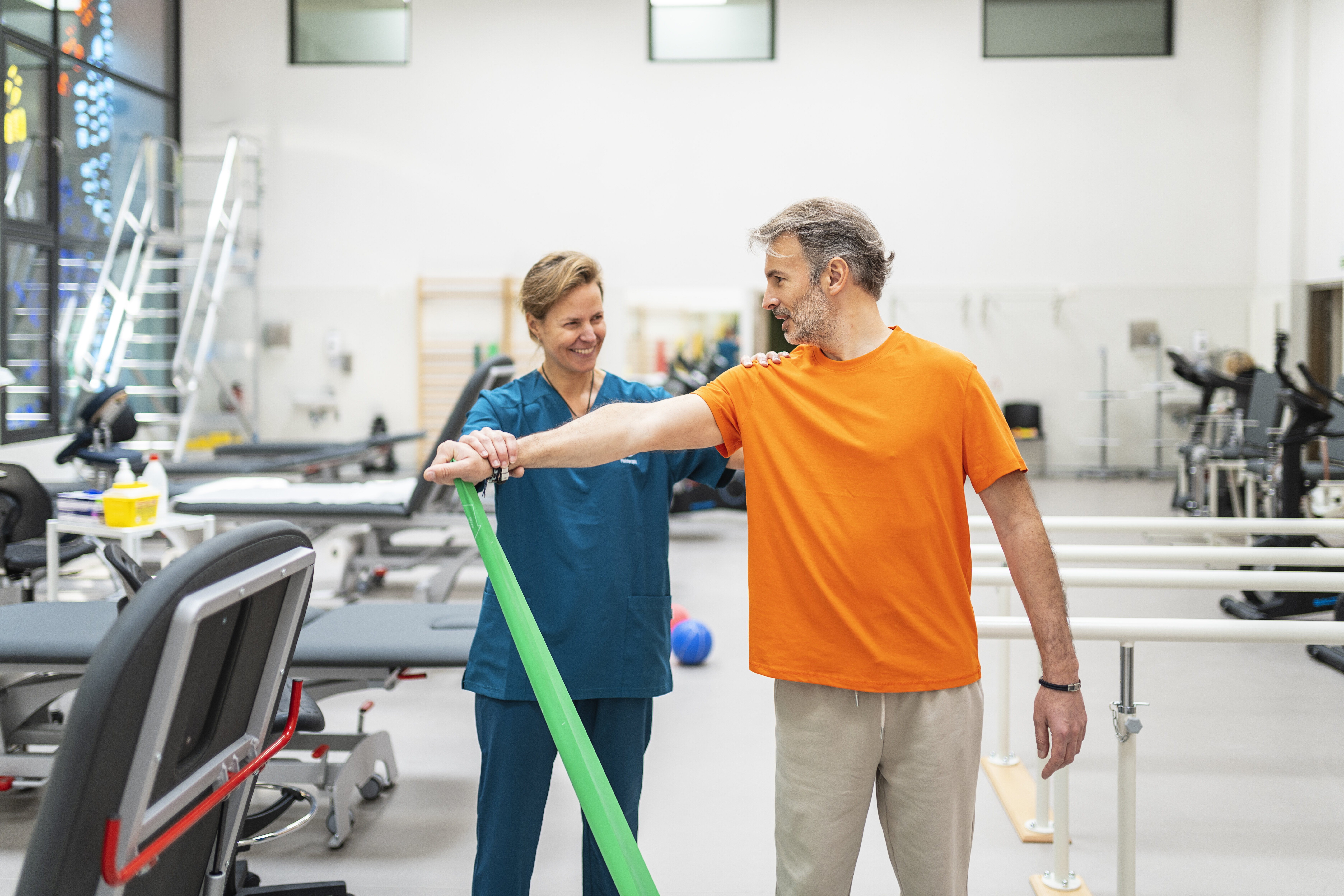 Physical therapist assists a man in an orange shirt with arm exercise using a green resistance band in a rehabilitation gym.