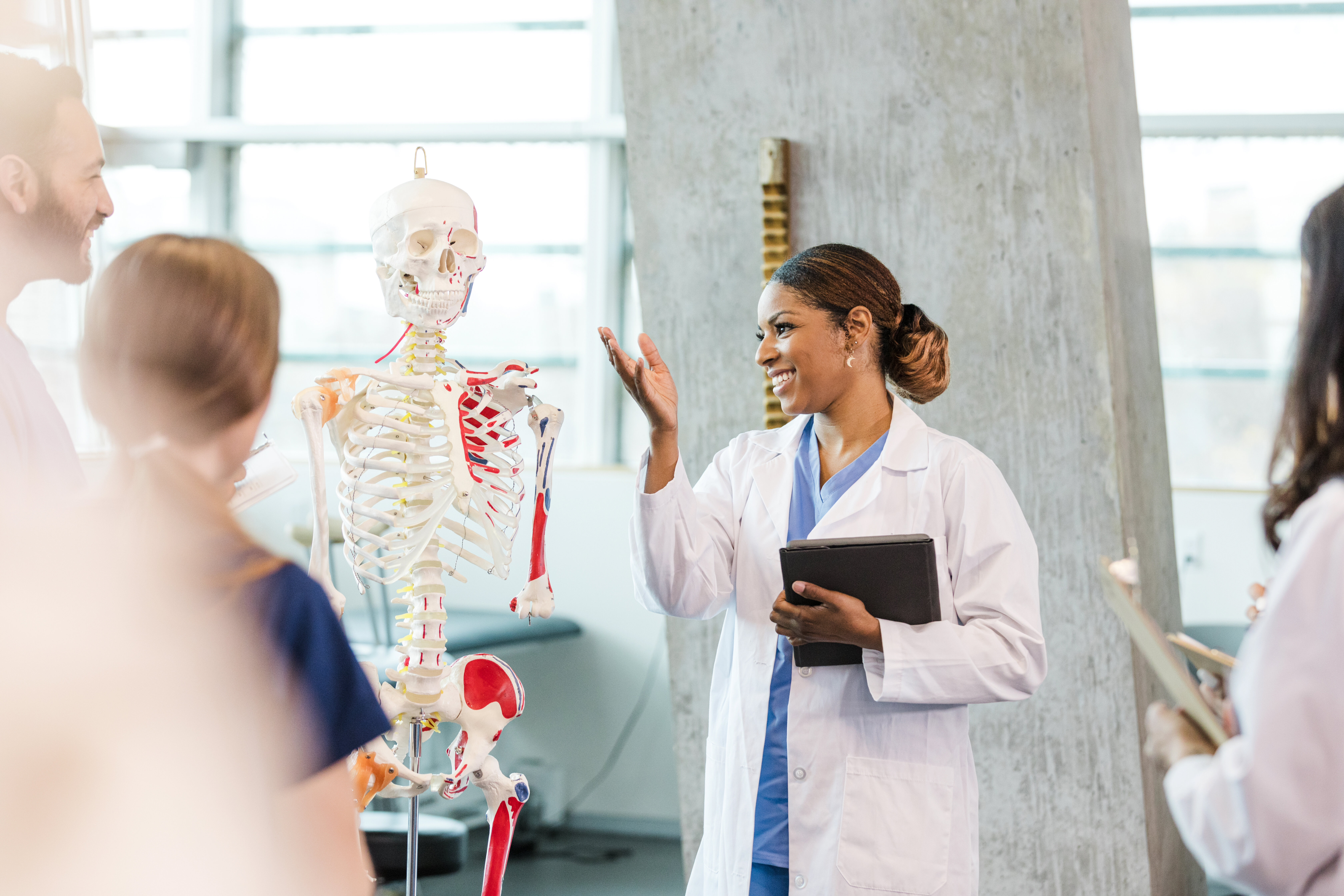 Medical instructor in white coat explaining human skeleton model to a group of students in a classroom.