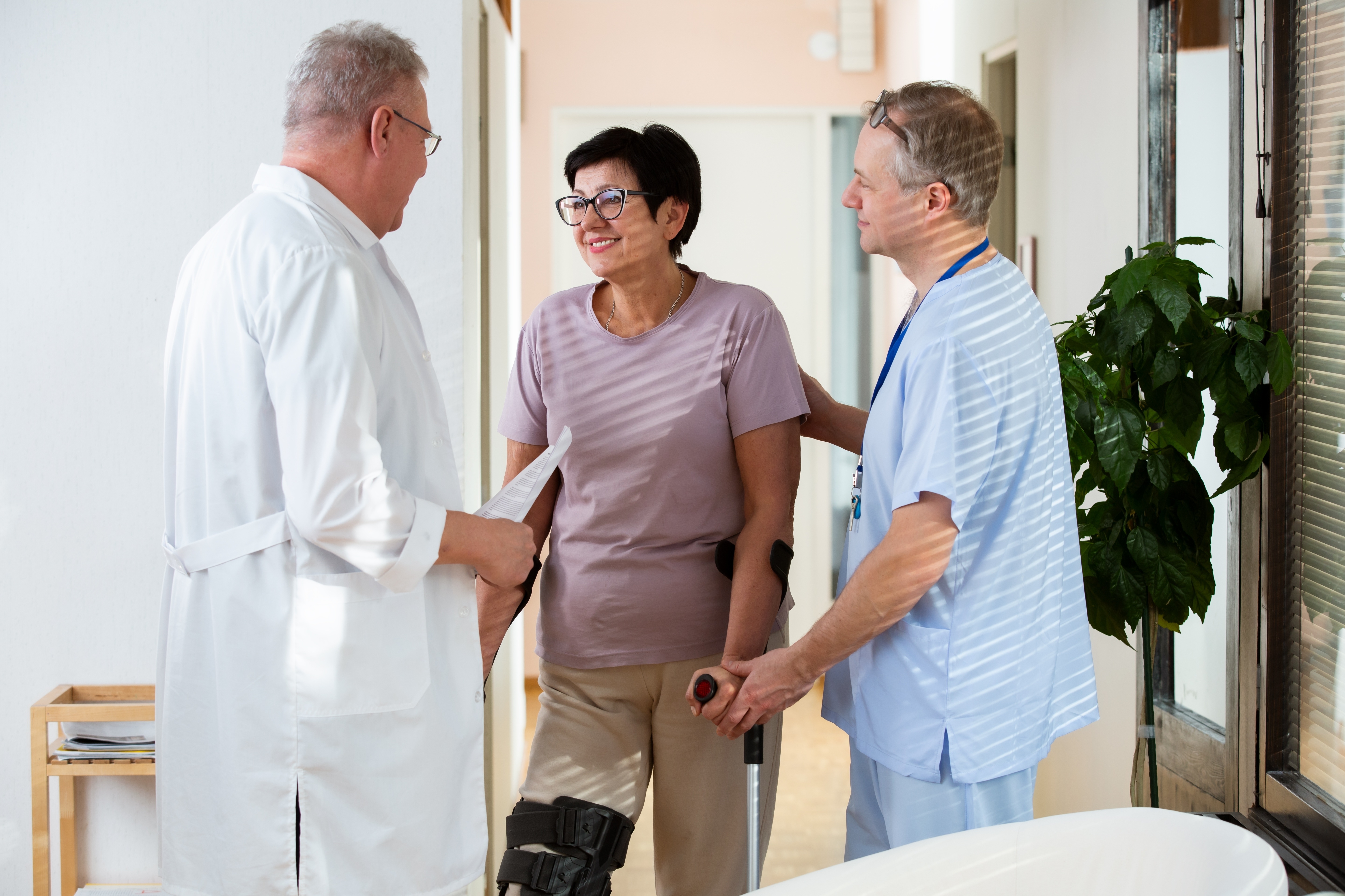 Female patient with leg brace and crutches talking to two male medical professionals in a clinic hallway.