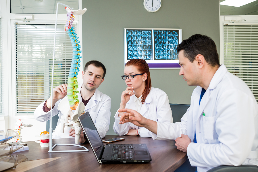 Three medical professionals in white coats discussing a colorful spine model while looking at a laptop in a clinical office.