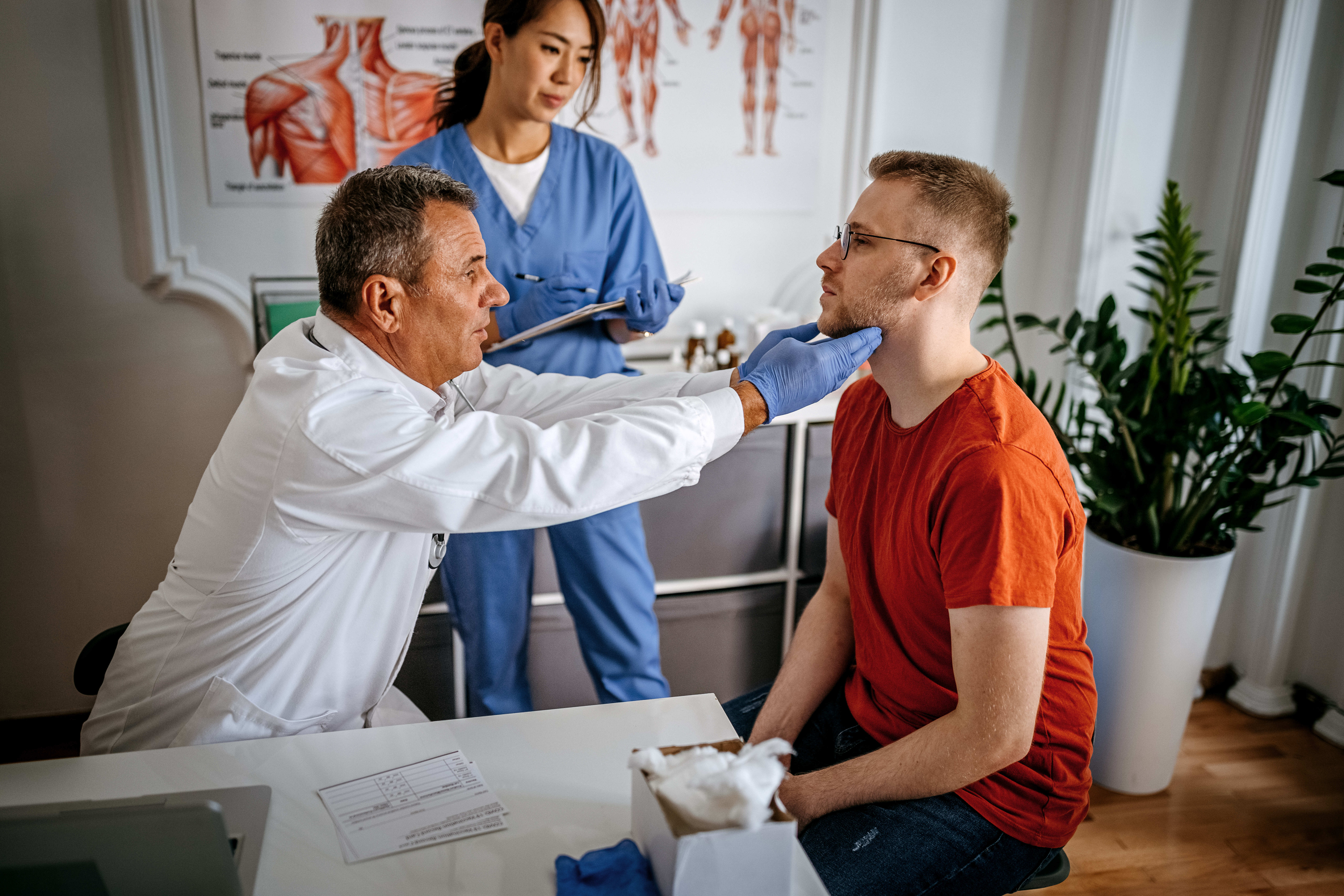 Doctor wearing gloves examining the throat of a seated male patient while a nurse stands behind taking notes.