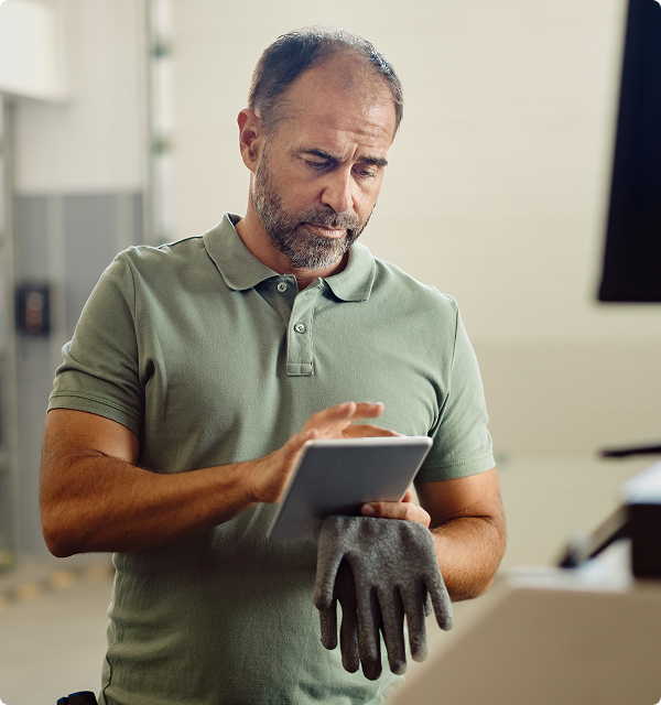 Man in green polo shirt holding a tablet and a pair of work gloves, looking at the screen in an industrial setting.