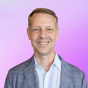 Smiling man with short light brown hair wearing a blue blazer and white shirt against a purple gradient background.