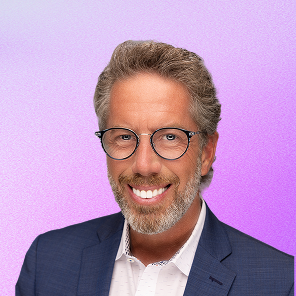 Smiling middle-aged man with glasses, gray beard, and curly hair wearing a navy blazer and white shirt against a purple gradient background.