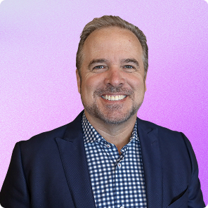 Smiling middle-aged man with short gray hair in a navy blazer and blue checkered shirt against a purple gradient background.