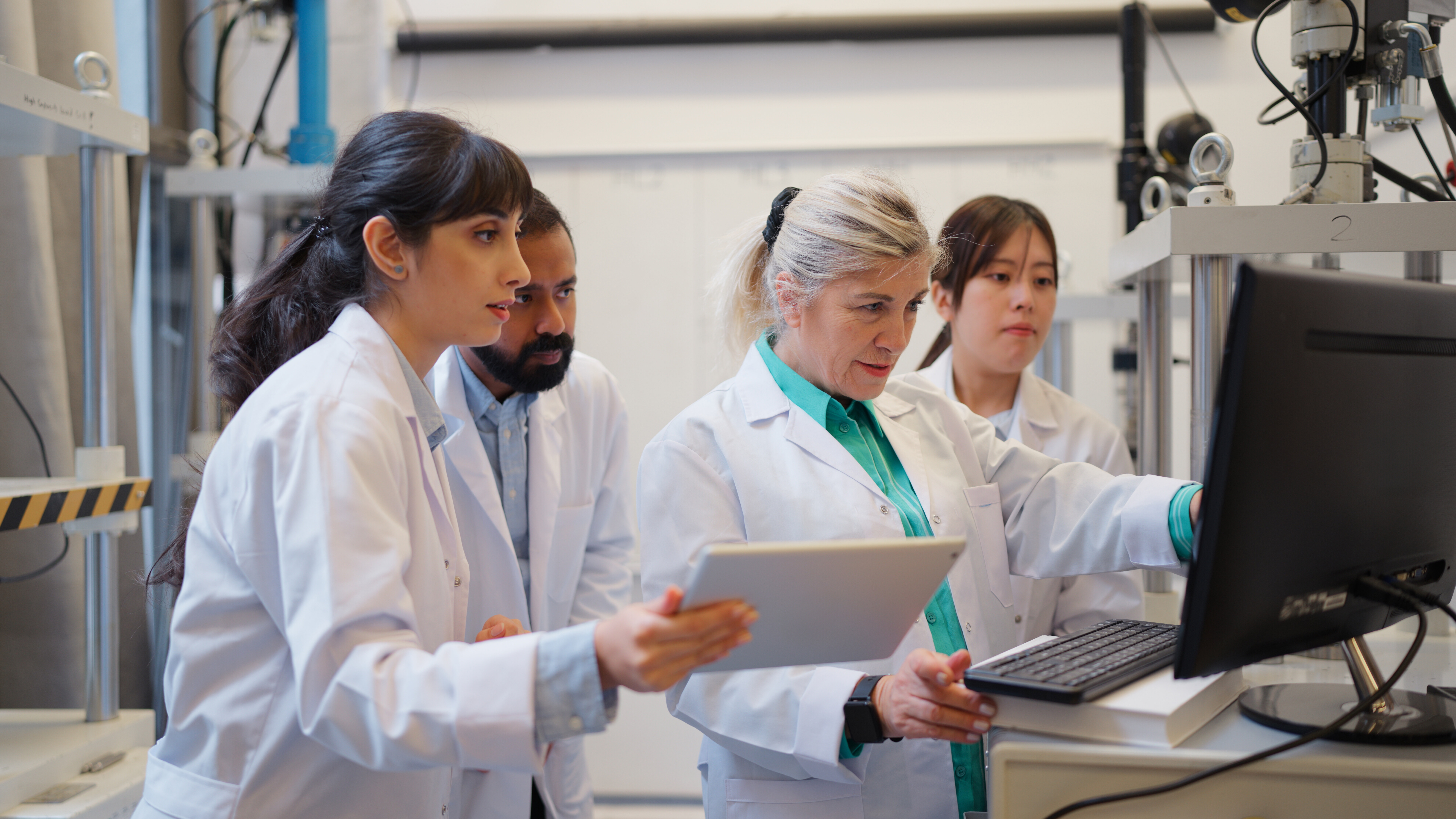 Four scientists in white lab coats collaborately looking at a computer screen in a laboratory.