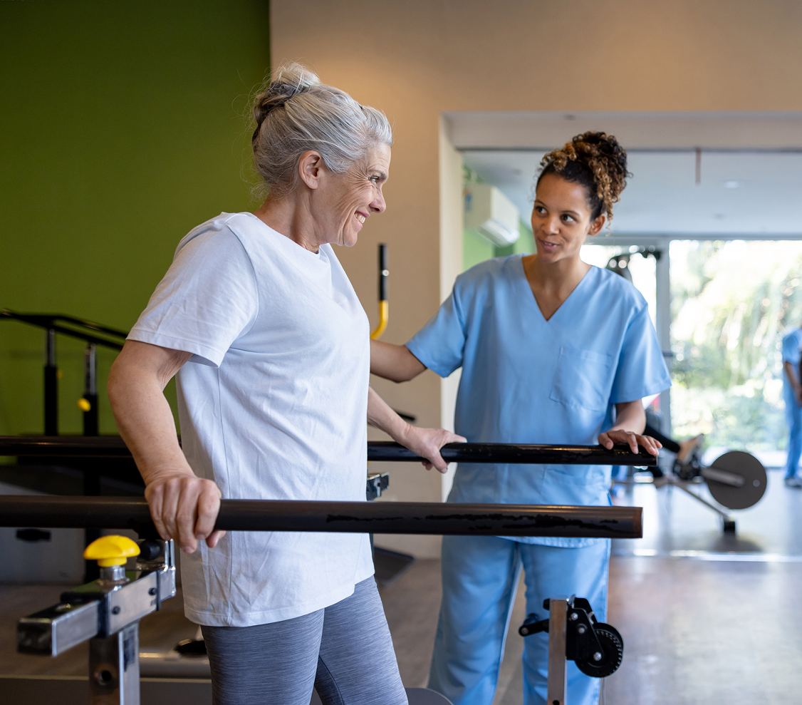 Older woman in white shirt using parallel bars for walking exercises assisted by a healthcare professional in blue scrubs.