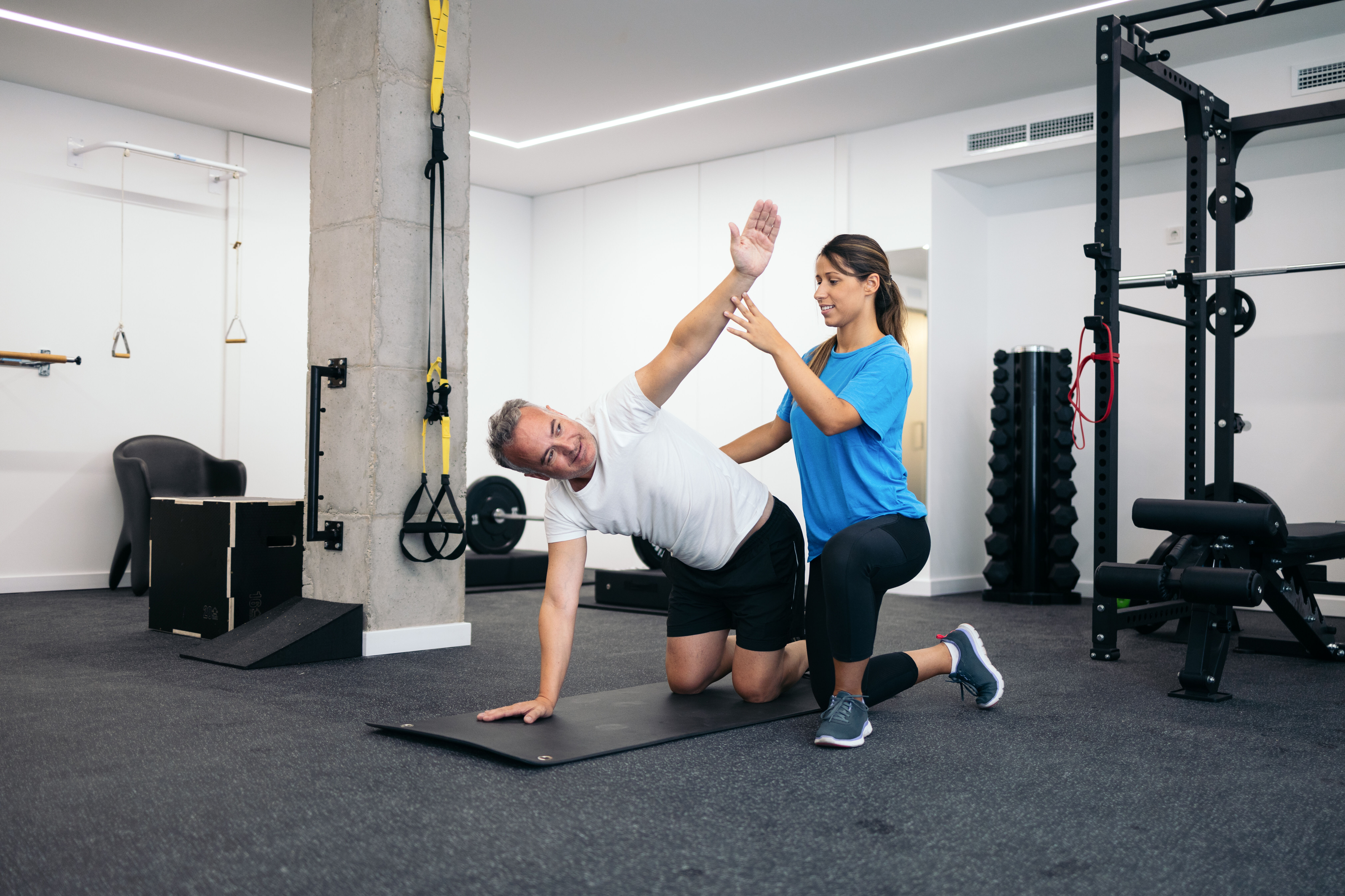 Fitness trainer assisting a man in a side stretch exercise on a mat in a gym.
