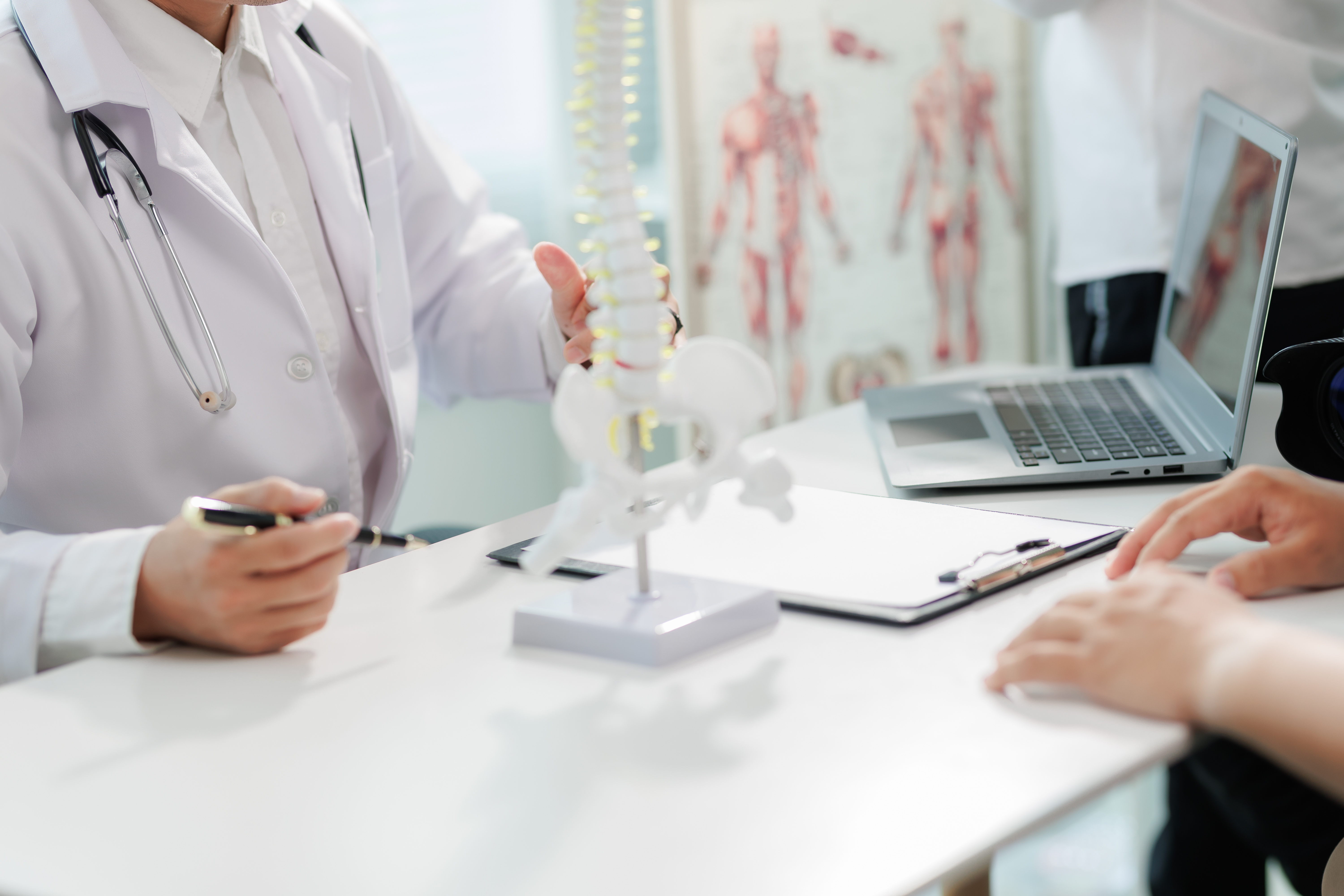 Doctor in white coat with stethoscope explaining spine model to patient at a desk with laptop and clipboard.