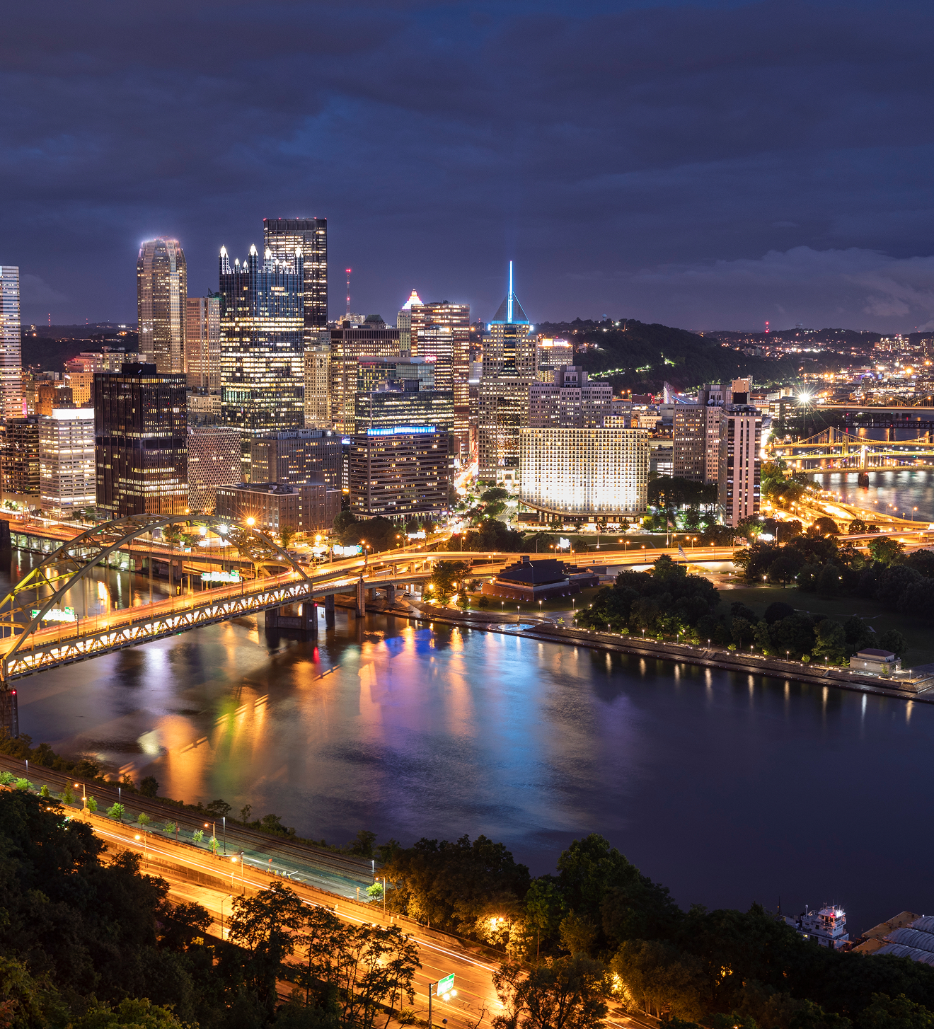 Night view of Pittsburgh skyline with illuminated buildings, bridges, and reflections on the river.
