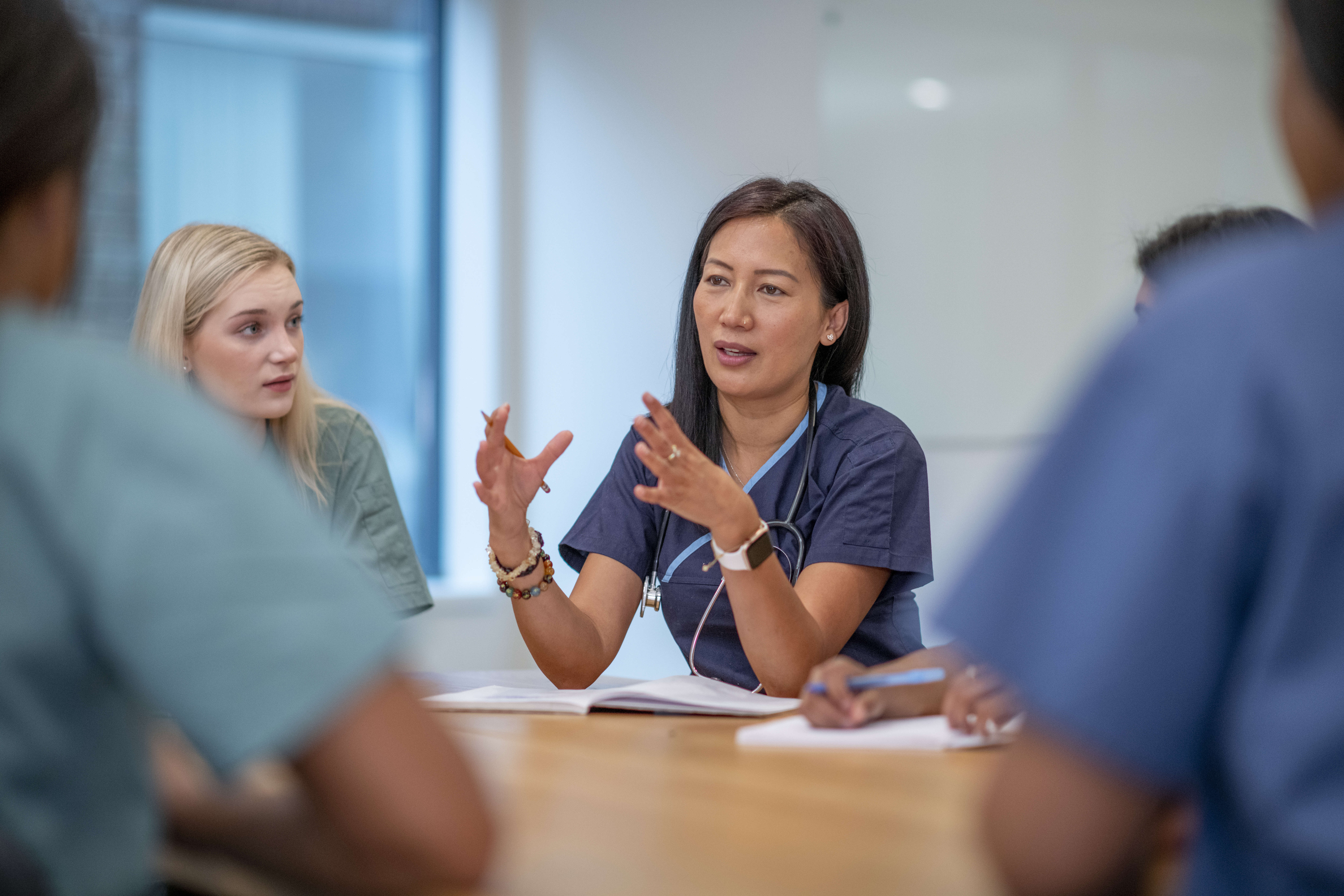 Female doctor speaking and gesturing with hands during a meeting with medical staff around a table.