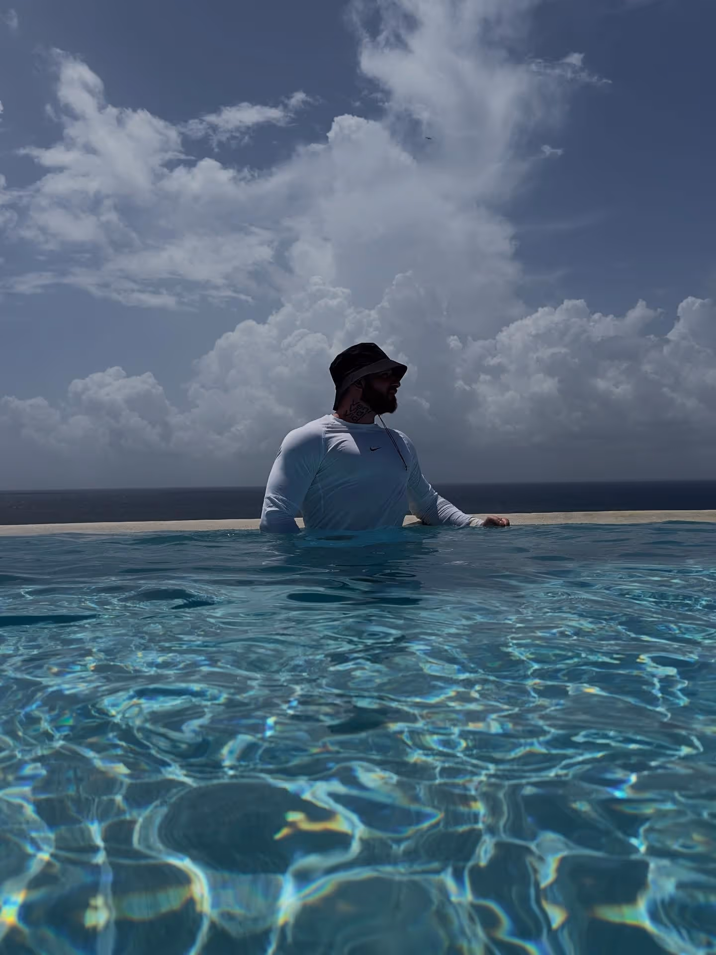 Portrait du coach Alexandre Millette : homme portant un chandail blanc à manches longues et un chapeau noir, debout dans une piscine sous un ciel nuageux – Coach Getfit, entraîneur privé à Québec.