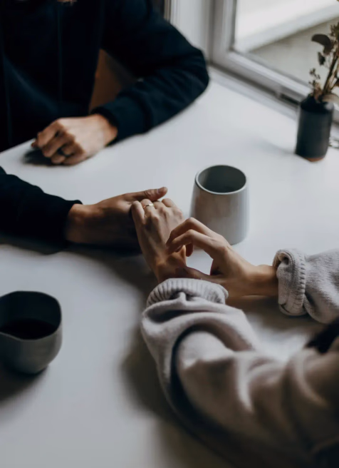 Two people holding hands across a white table with ceramic cups and a small plant near a window.