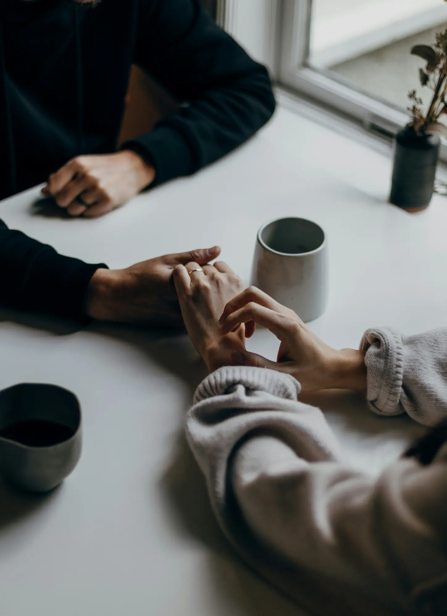 Two people holding hands across a white table with ceramic cups and a small plant near a window.