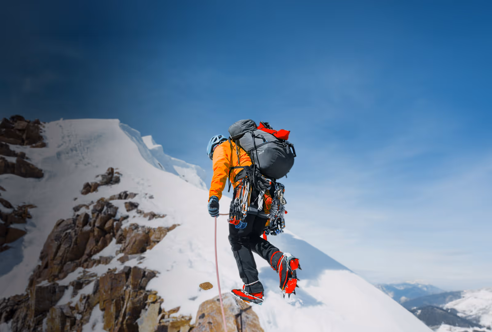 Mountaineer in orange jacket climbing snowy mountain ridge with climbing gear and crampons under clear blue sky.
