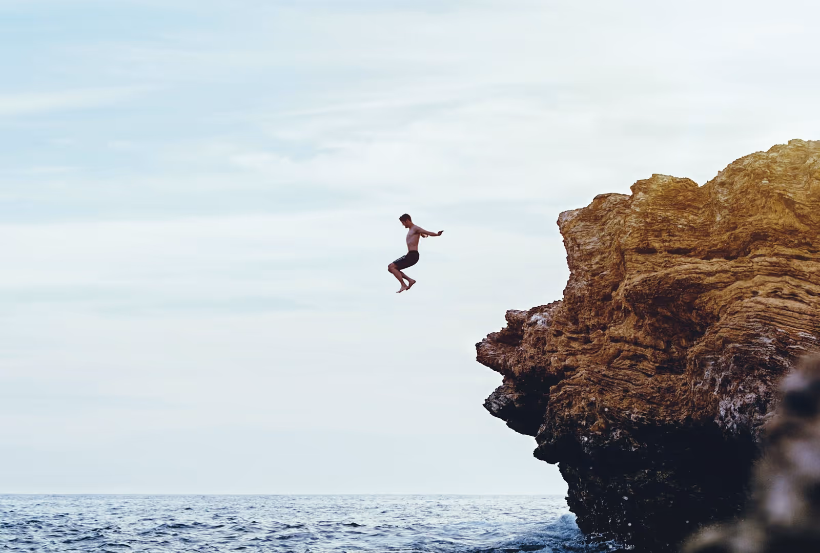 Person jumping off a rocky cliff into the ocean under a cloudy sky.