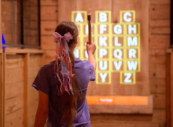 Person with long hair tied with a large pink ribbon holding an axe, pointing at an illuminated grid of alphabet letters on a wooden wall.