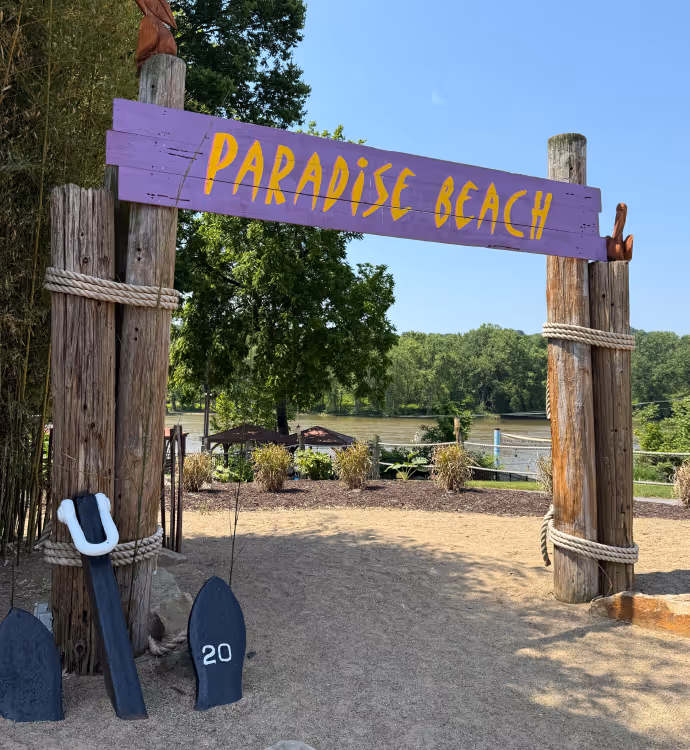 Wooden archway with a purple sign reading 'PARADISE BEACH' in yellow letters, with trees and a river visible in the background.
