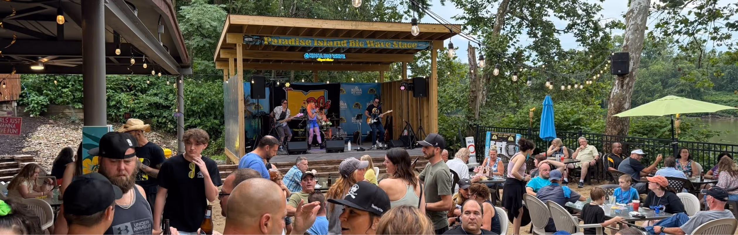 Outdoor music event with a band performing on a wooden stage labeled 'Paradise Island Big Wave Stage' surrounded by a crowd seated at tables under string lights and umbrellas.