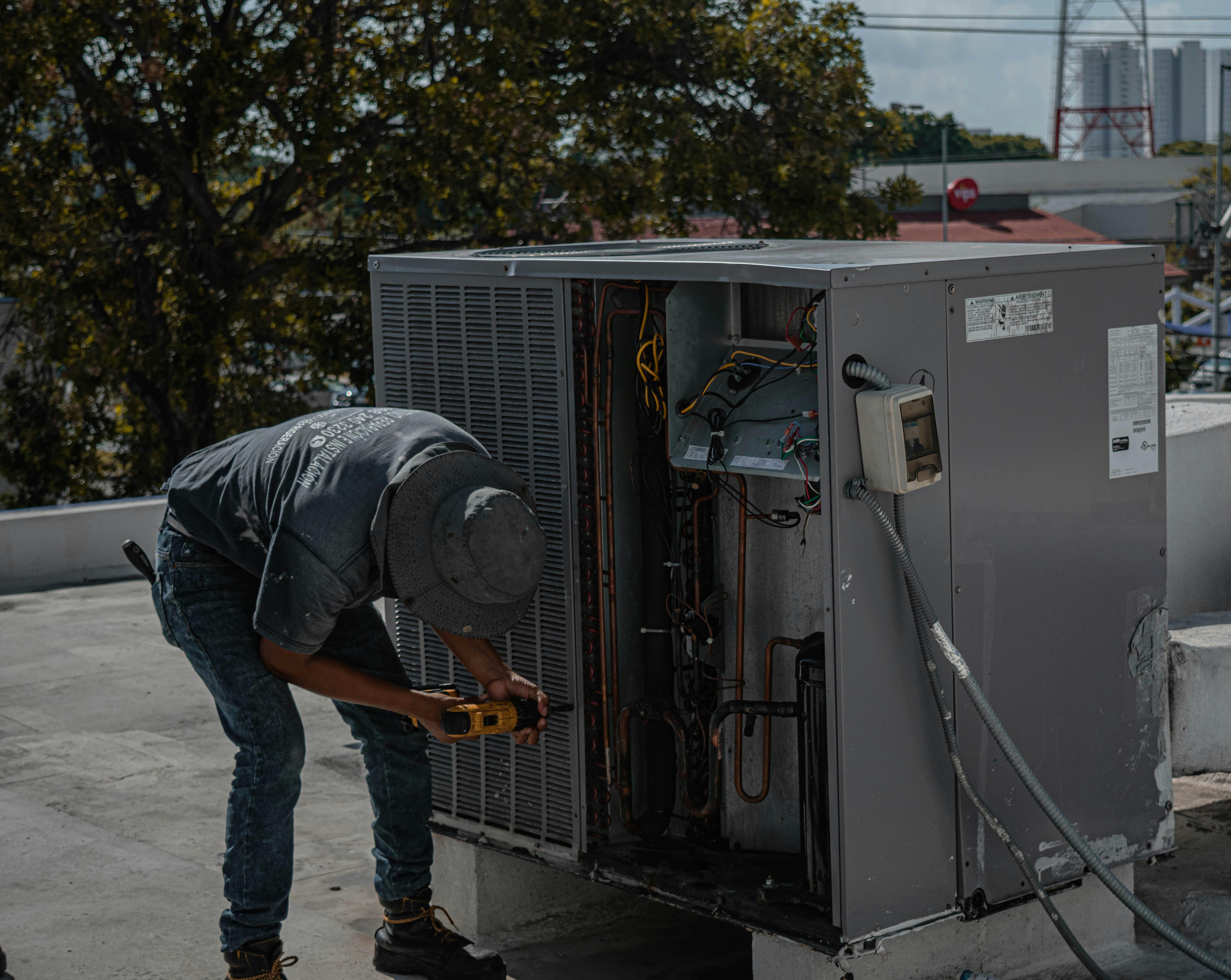 Technician in a hat and work boots repairing an HVAC unit on a rooftop with a power drill.