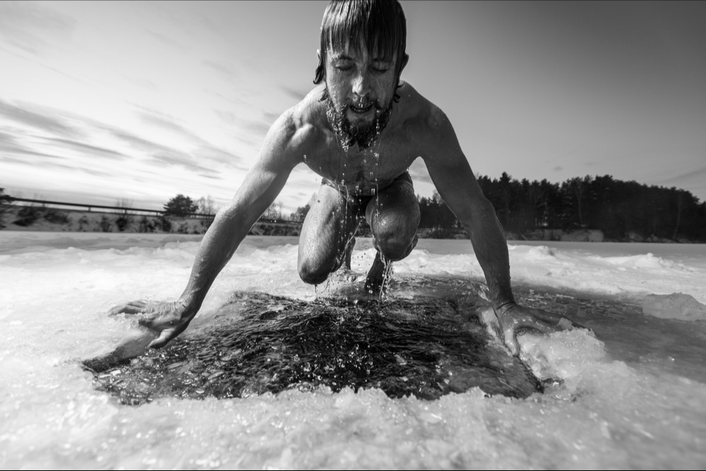Man emerging from a rectangular hole cut in frozen ice on a snowy landscape at sunset.