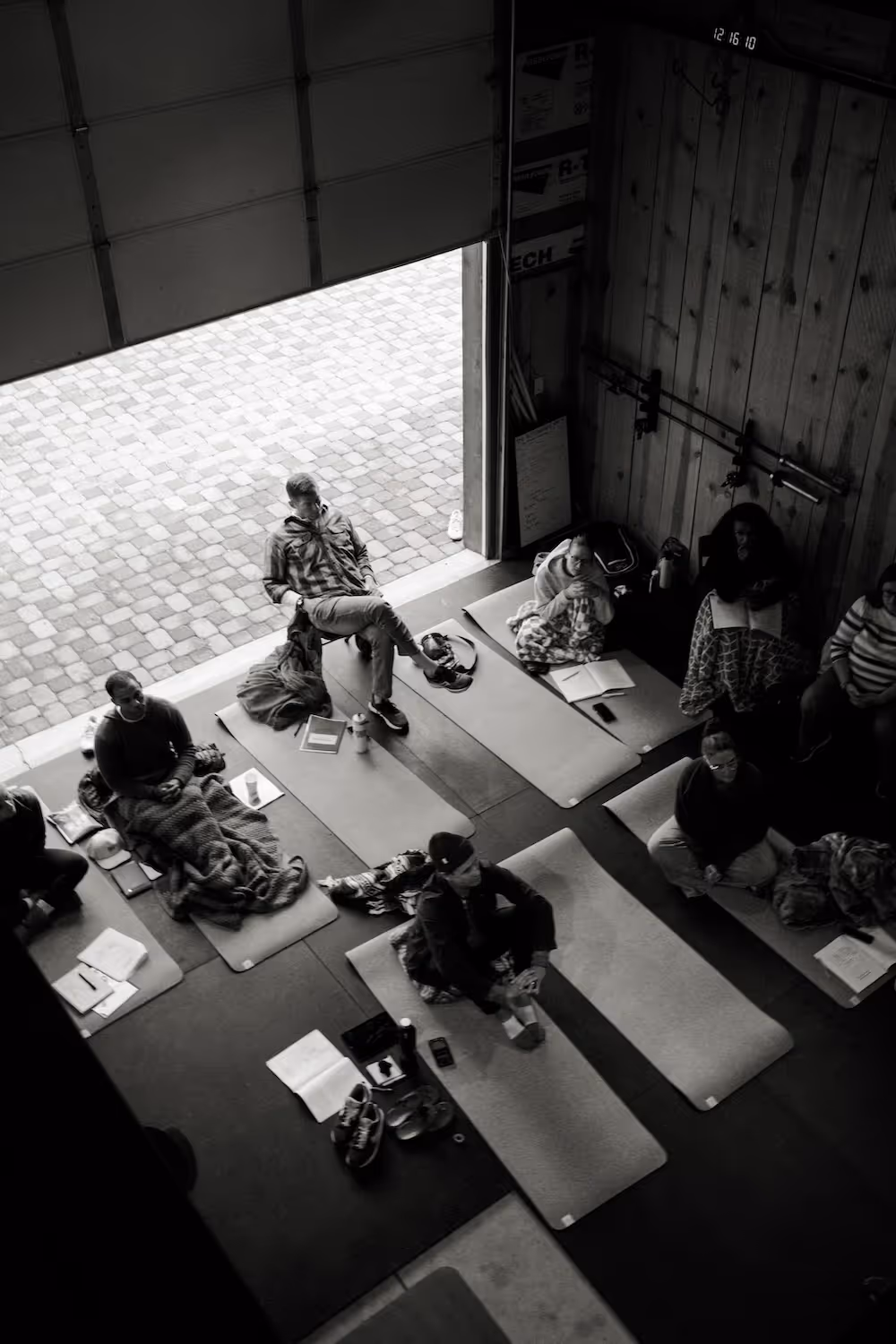 People sitting on yoga mats inside a dimly lit room with wooden walls, some reading books and others wrapped in blankets.
