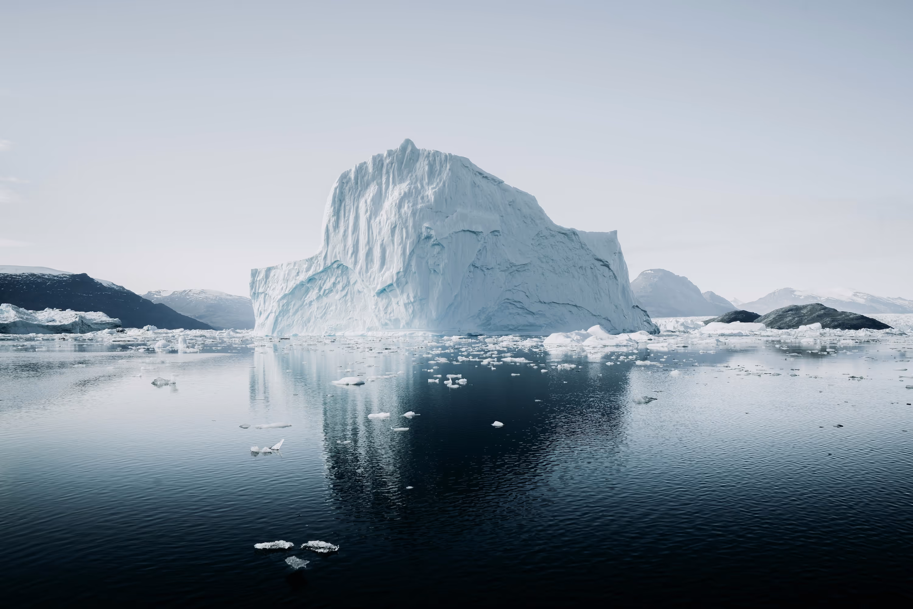 Large iceberg floating in calm Arctic water with smaller ice chunks and distant snow-covered mountains.