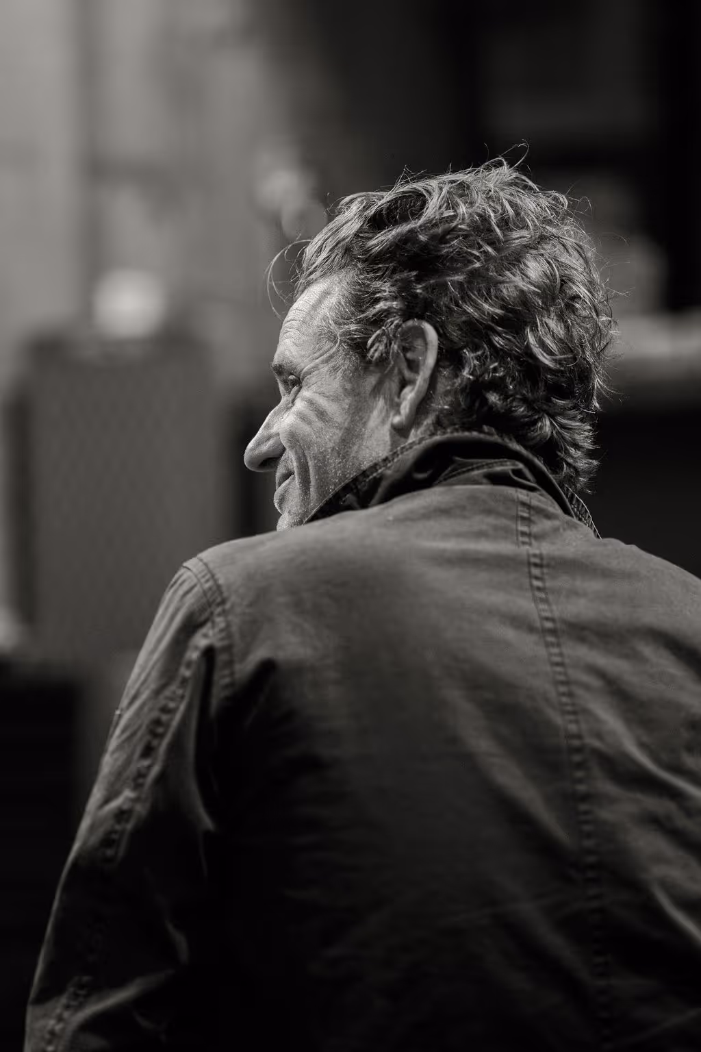 Black and white photo of a man with curly hair smiling, seen from behind and slightly to the side.