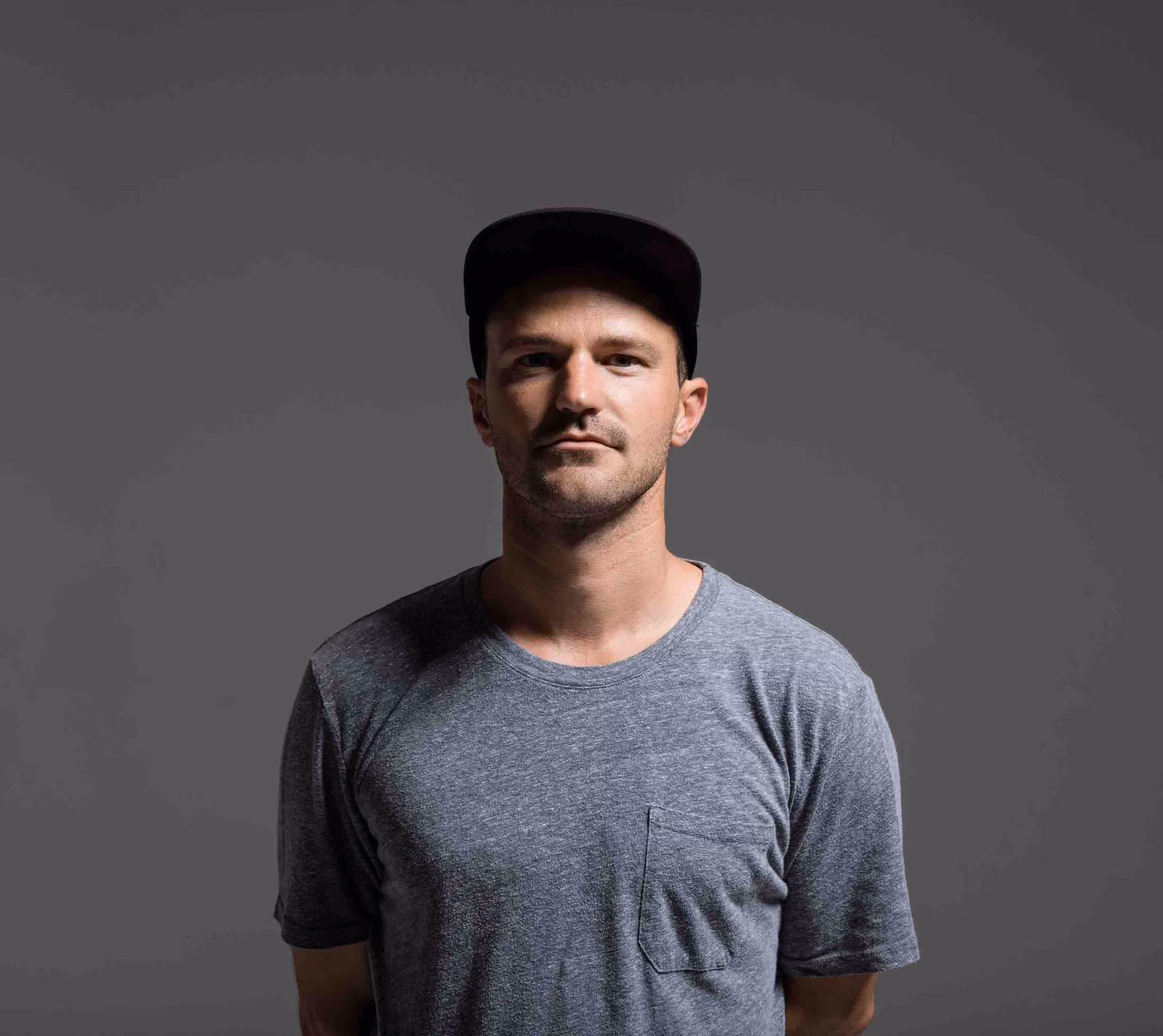 Man wearing a gray t-shirt and black cap, standing against a plain gray background with a neutral expression.