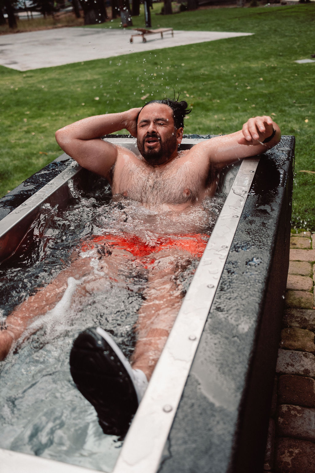 Man in orange swim trunks sitting in an outdoor ice bath with water splashing around him.