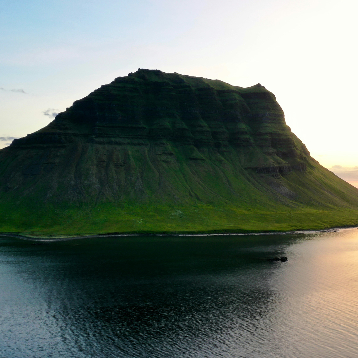 Steep green mountain rising beside a calm water body at sunset under a clear sky.