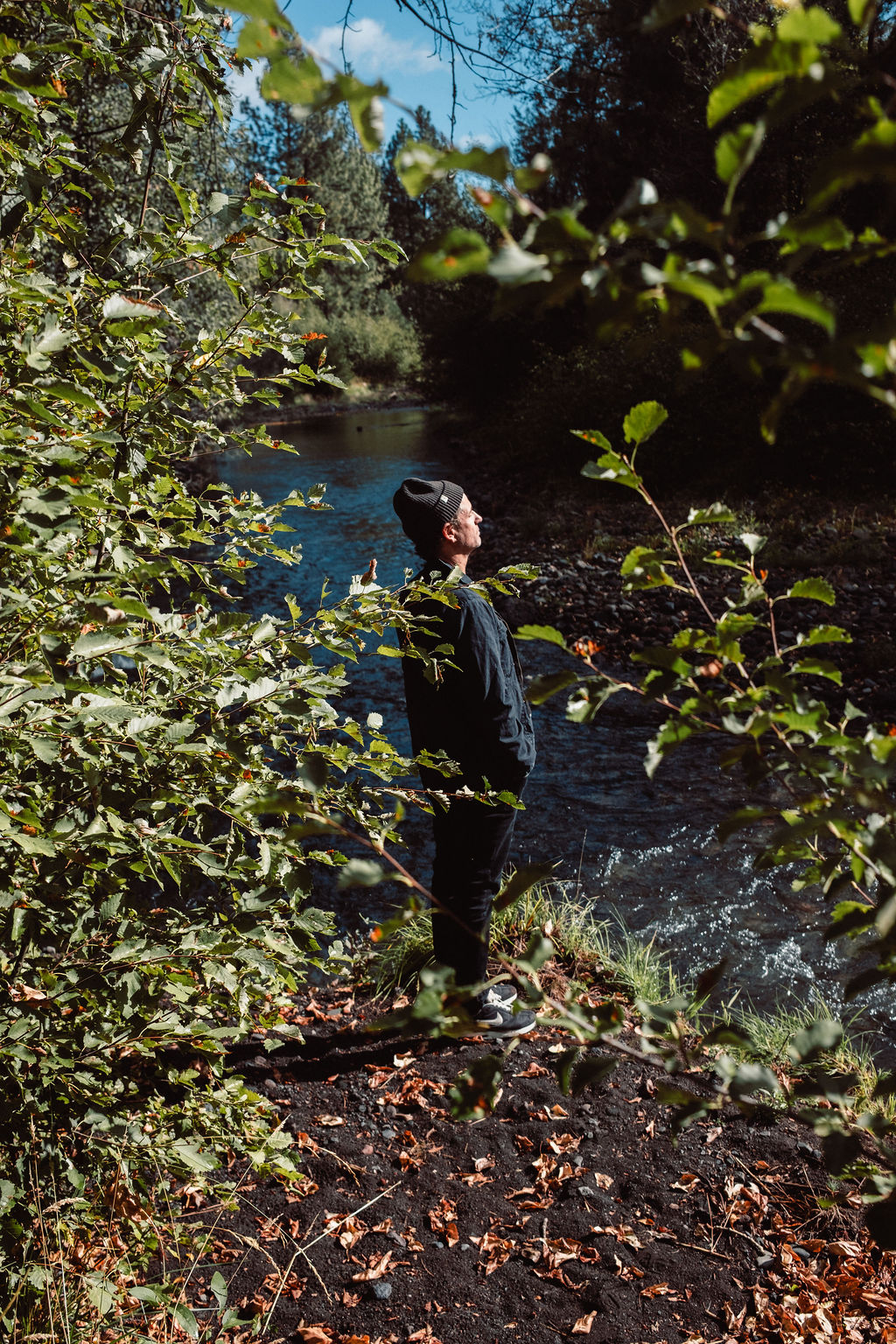 Man in a black jacket and beanie standing near a riverbank surrounded by green leafy trees and autumn leaves.