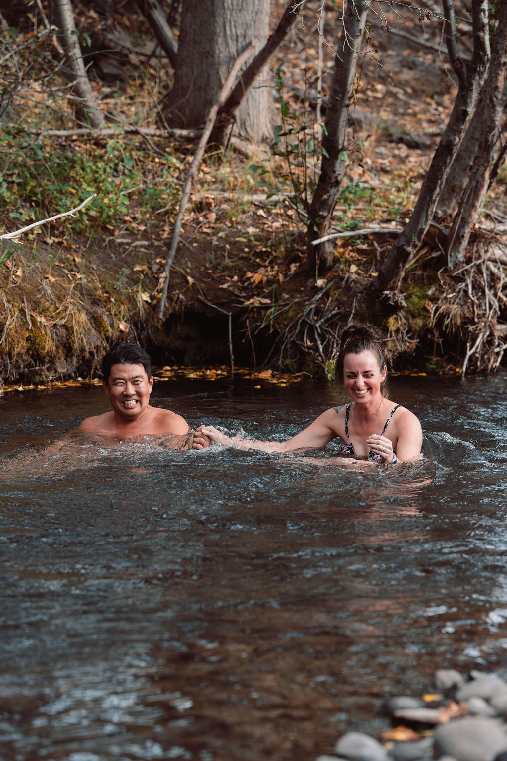Smiling man and woman holding hands while standing in a shallow river near a wooded riverbank.