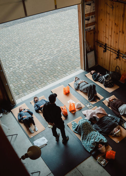 Six people lying on yoga mats indoors near an open garage door, with one standing person overseeing them.