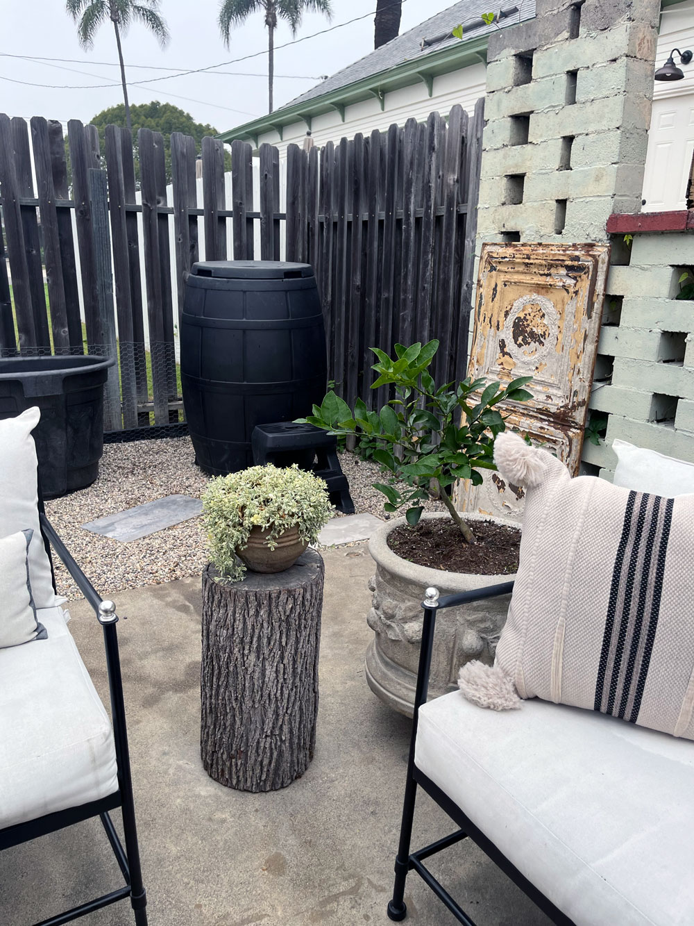 Outdoor patio area with two cushioned chairs, a small tree stump table holding a potted plant, and a larger potted plant near a weathered wooden panel and black fence.