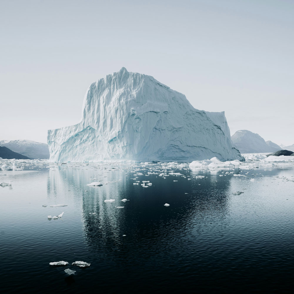 Large iceberg floating on calm dark water with smaller ice chunks and distant snowy mountains under a pale sky.