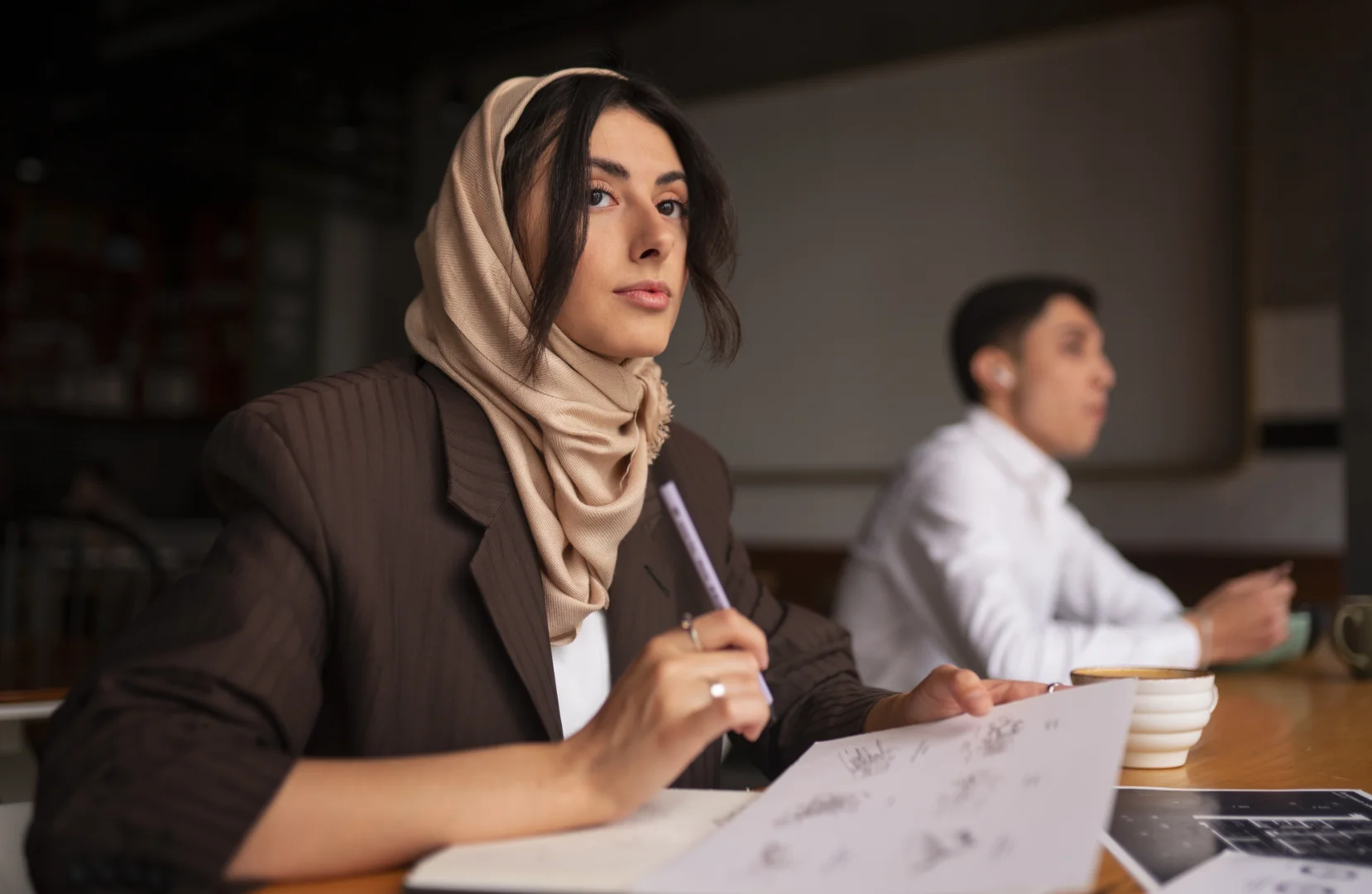 Young woman wearing a beige hijab and brown jacket sitting at a table, holding a pen and a paper with sketches.