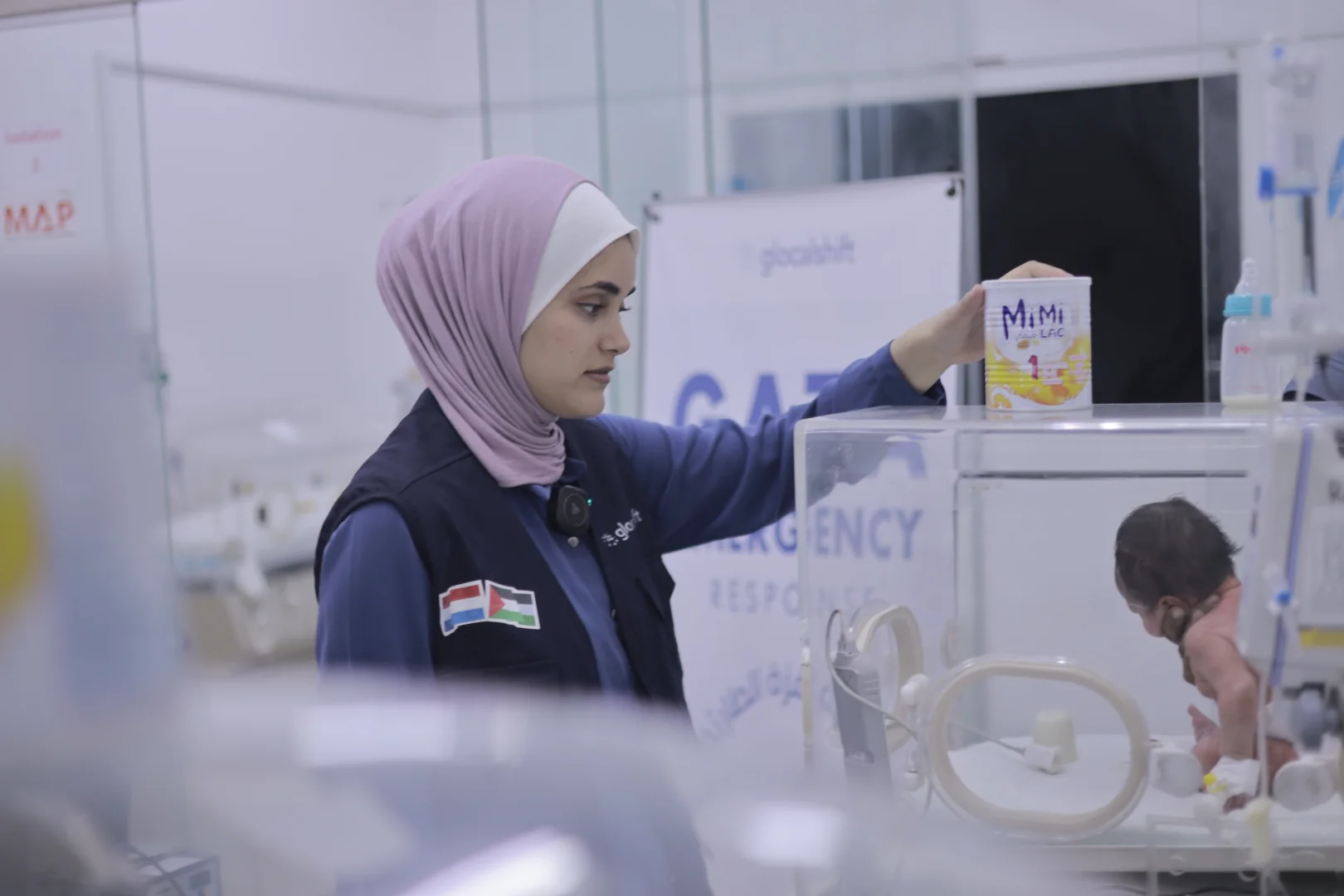A woman wearing a pink hijab and navy vest with flags tending to a baby in a hospital incubator in Gaza