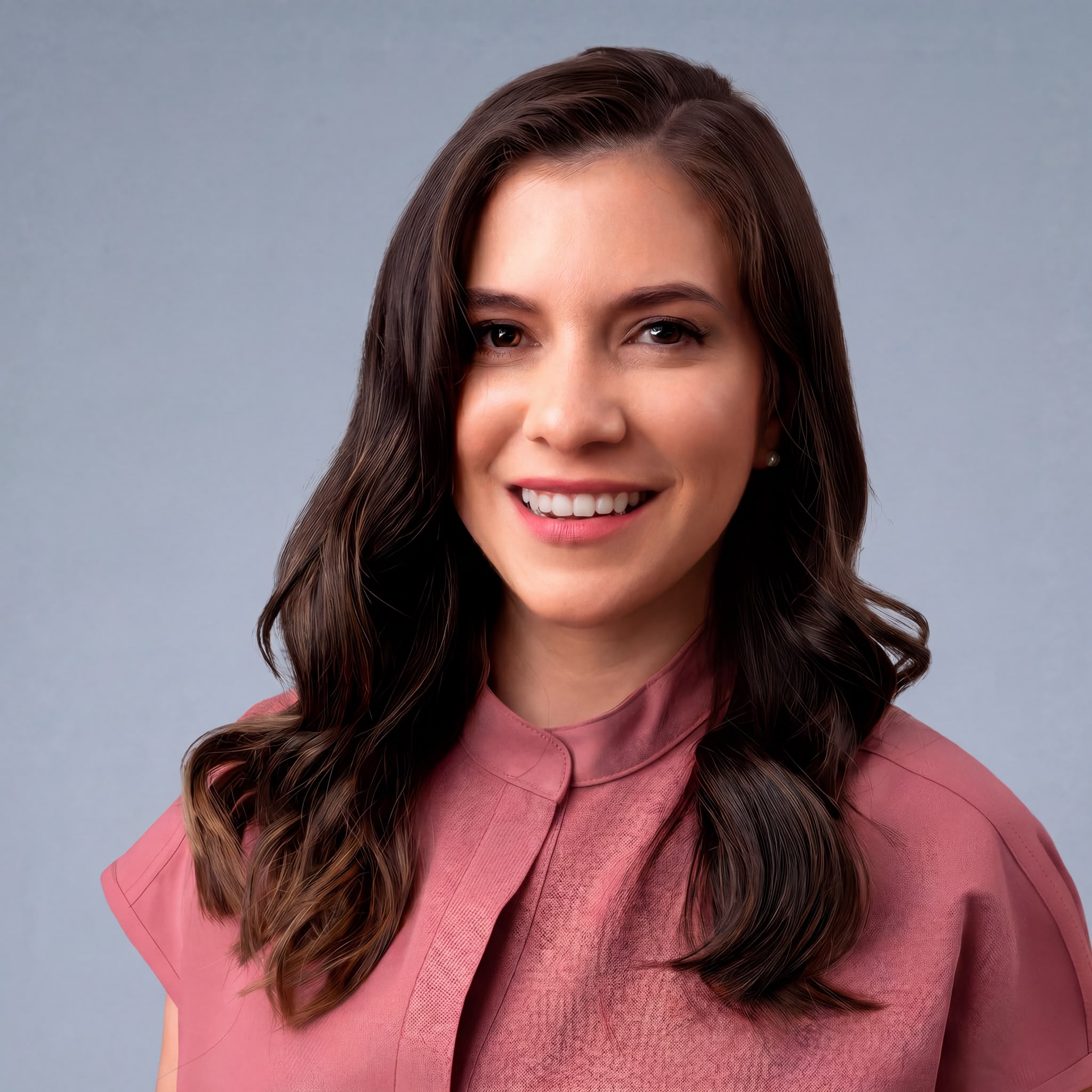 Smiling woman with wavy brown hair wearing a pink blouse against a light gray background.