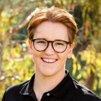 Smiling person with short reddish hair and glasses wearing a black collared shirt outdoors with blurred greenery background.