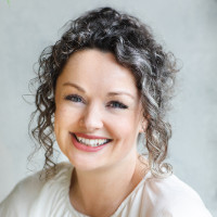 Smiling woman with curly dark hair wearing a white top against a light gray background.