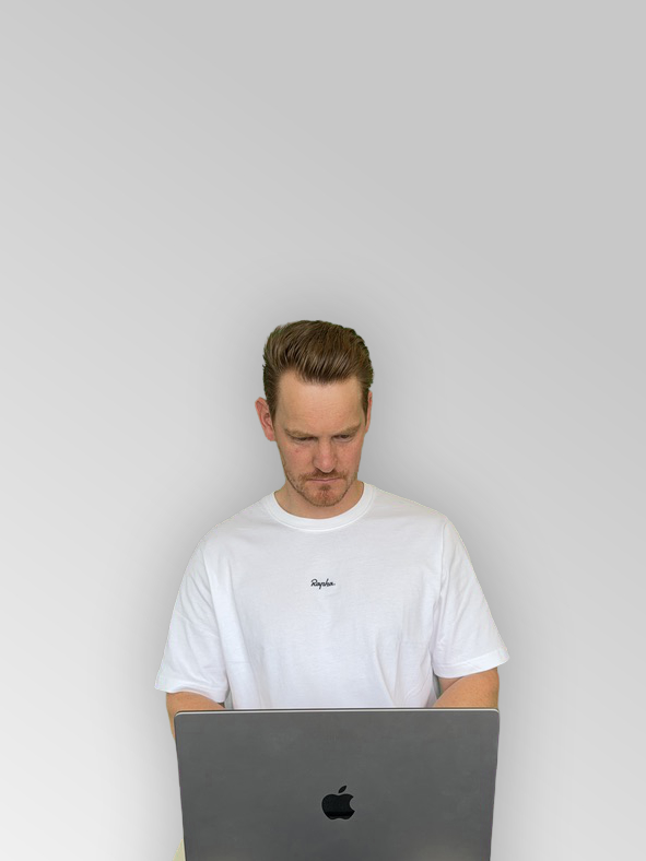 Me with short red hair wearing a white t-shirt working on an Apple laptop against a plain gray background.