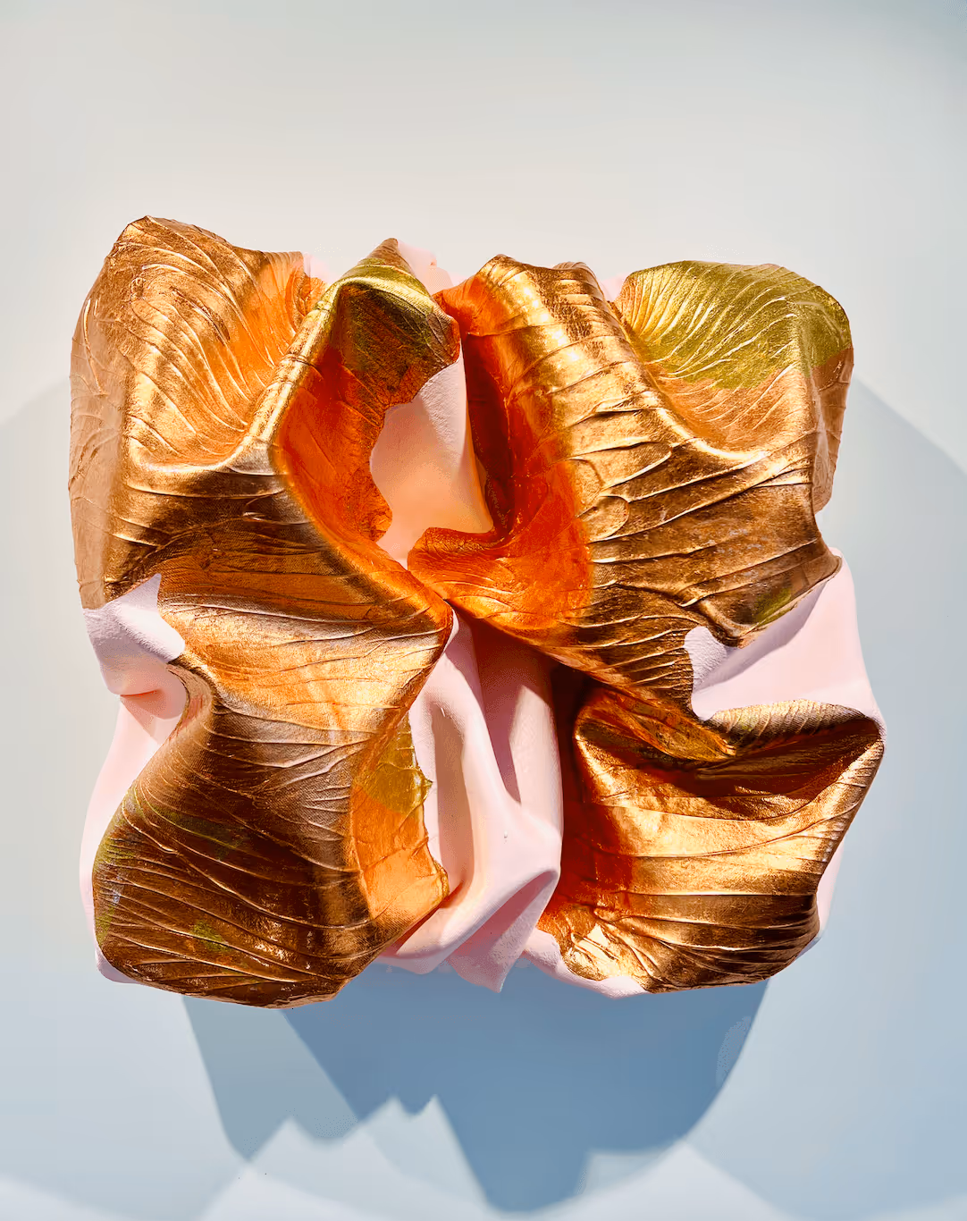 Abstract sculpture with folded, textured metallic gold and soft pink surfaces casting shadow on a light background.