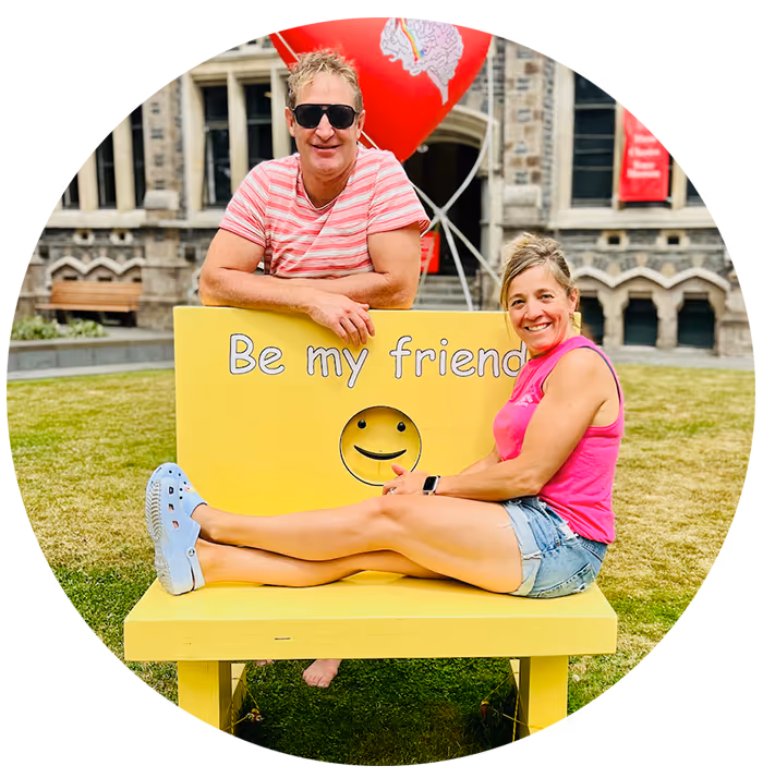 Ben Reid in sunglasses and pink striped shirt leans on a yellow bench with a smiley face and the text 'Be my friend' as a Olivia Reid in a pink tank top and denim shorts sits on the bench outdoors.