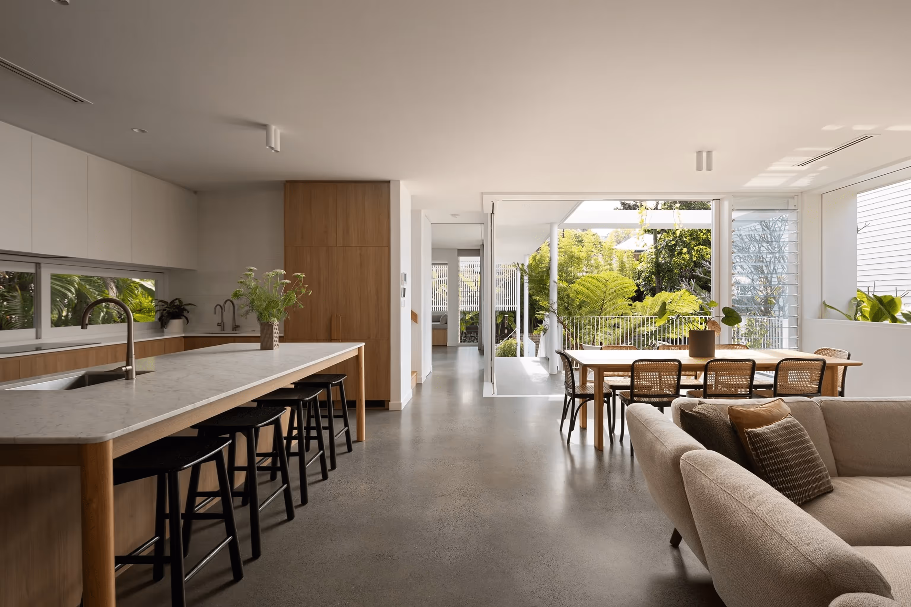 Modern open-plan kitchen and living area with wood and white finishes, large island seating, dining table, and sliding glass doors opening to a lush garden terrace.