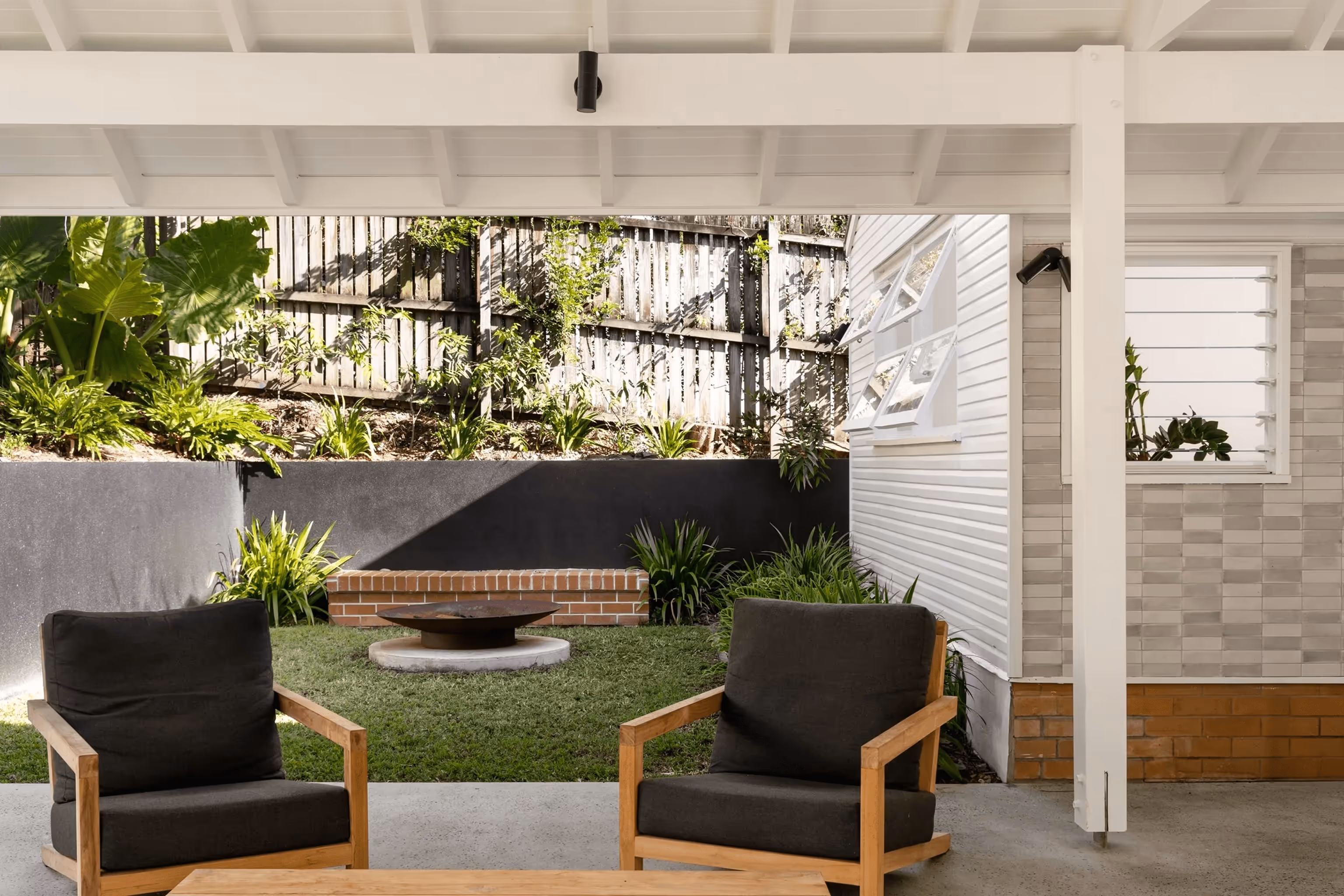 Covered outdoor patio with wooden chairs, a coffee table, and a view of a small garden featuring a fire pit and retaining wall.