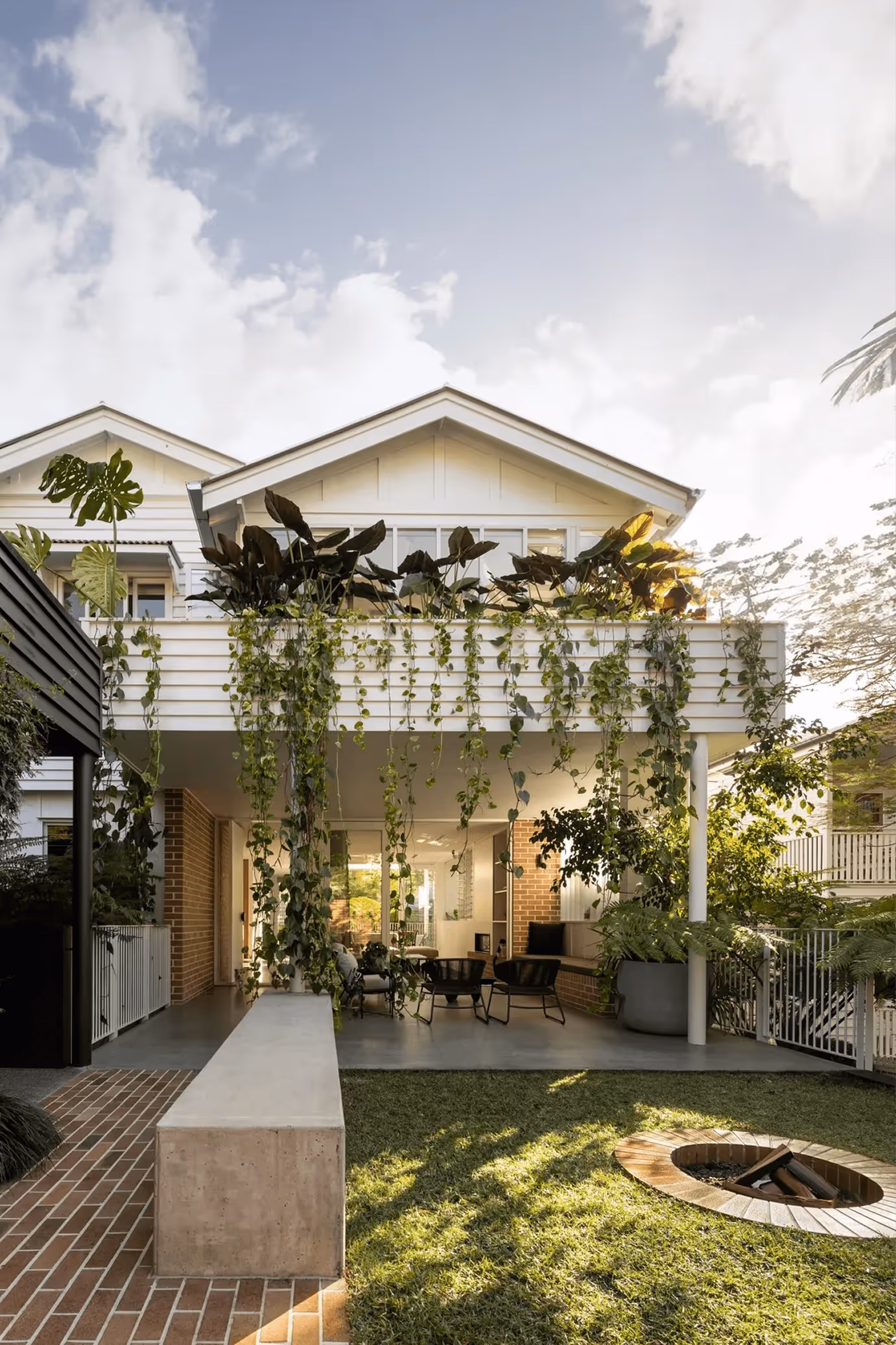 Two-story home with a leafy balcony overflowing with hanging plants, an open patio below, and a backyard featuring a fire pit at sunset.