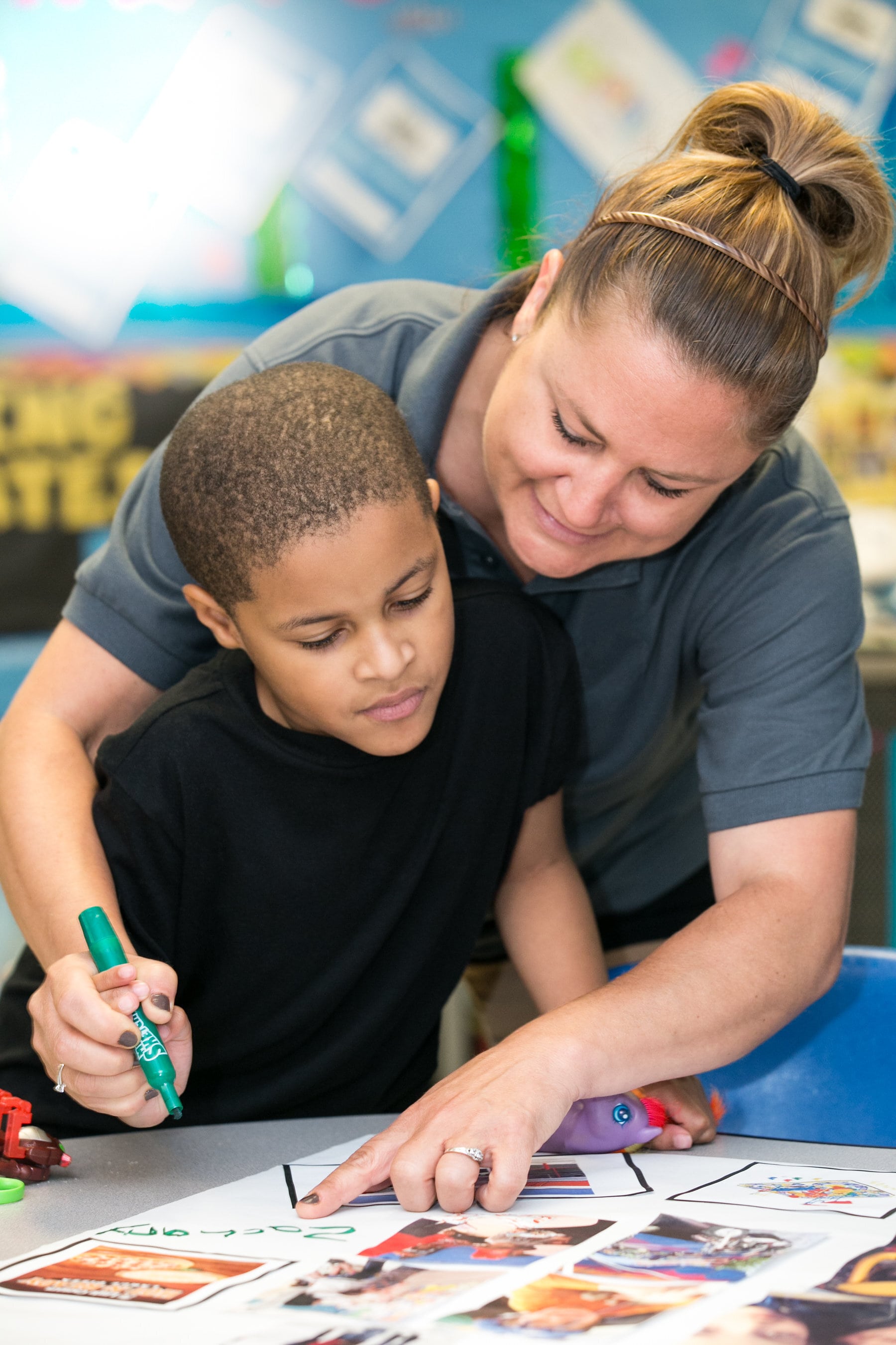 An adult helps a young boy with a worksheet at a table, guiding his hand as he writes with a green marker in a classroom setting.